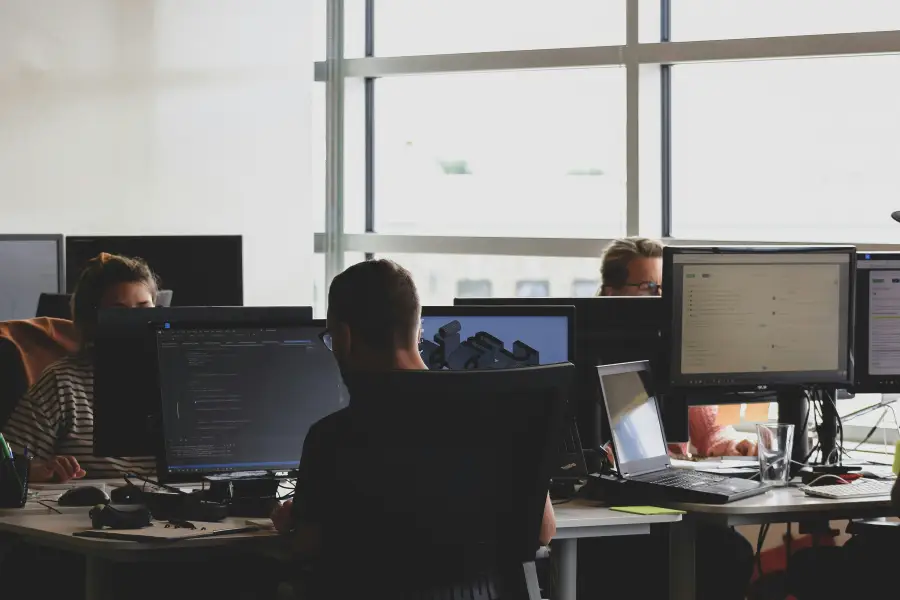 Tech-business professionals working at multi-monitor desks with code and 3D design software in a modern open office.