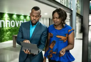Two tech-business professionals walk through a modern office lobby; reviewing data on a tablet together.